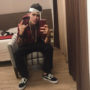 mc kevinho em selfie no espelho, sentado, usando corrente de ouro, bandana branca e jaqueta vinho