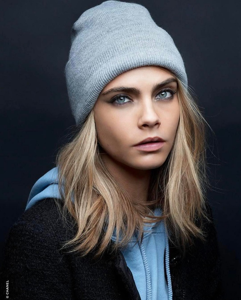 Atriz Cara Delevingne usando roupa de frio e touca, em fundo cinza
