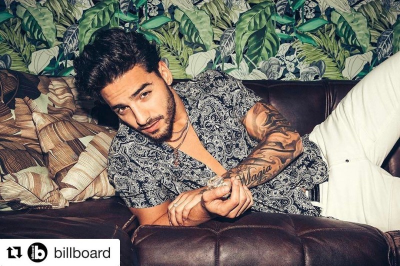 Maluma deitado, olhando para a câmera, usando camisa estampada preta e branca, em fundo estampado de folhas