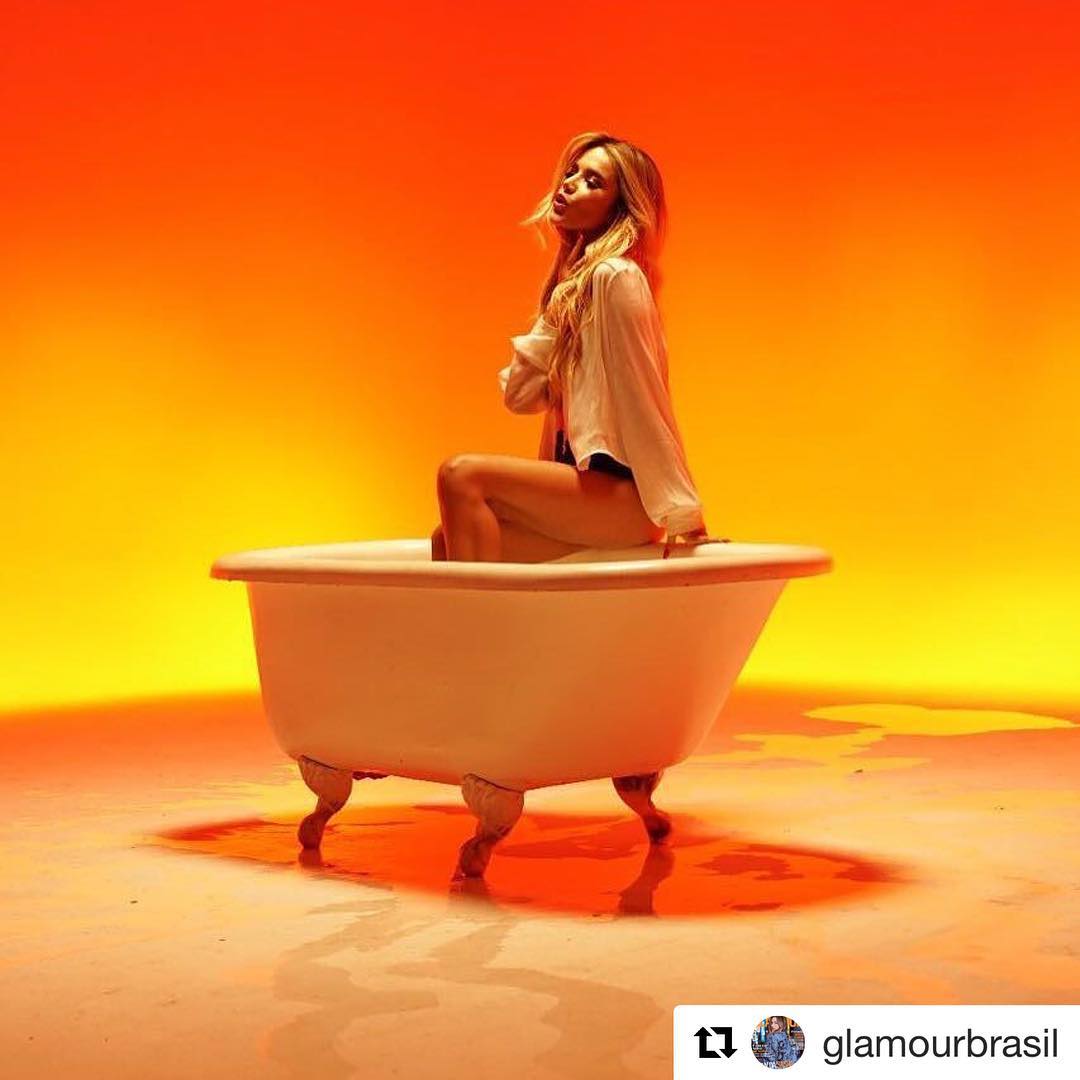 Manu Gavassi em clipe da música Hipnose, sentada em uma banheira branca, usando roupas brancas, em um fundo amarelo