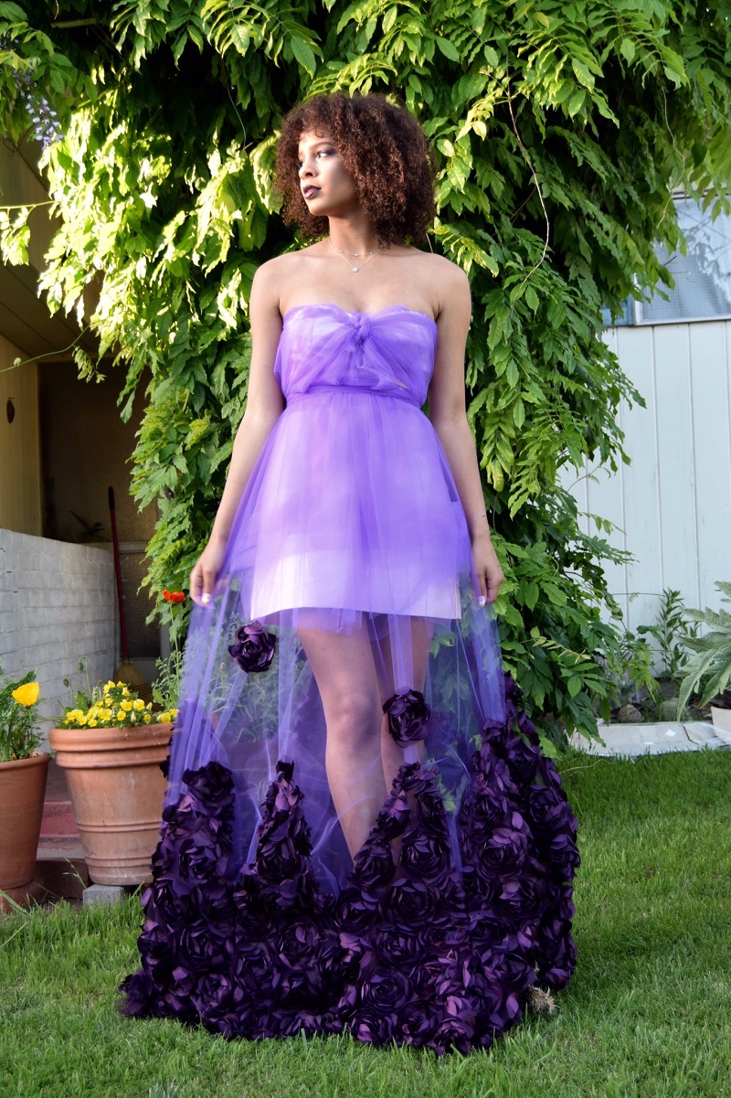 menina usa vestido roxo de tule, com flores na barra, em meio a um jardim