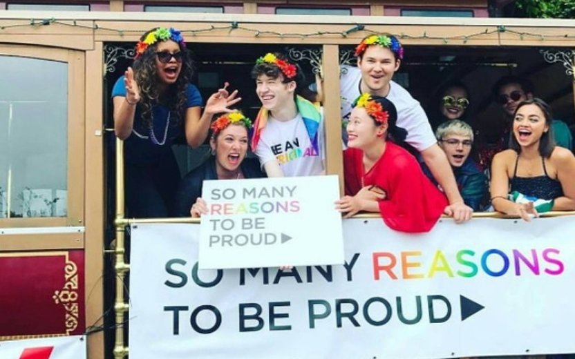 elenco de 13 reasons why segurando plaquinha que diz "muitas razões para se orgulhar", usando acessórios com as cores do arco íris