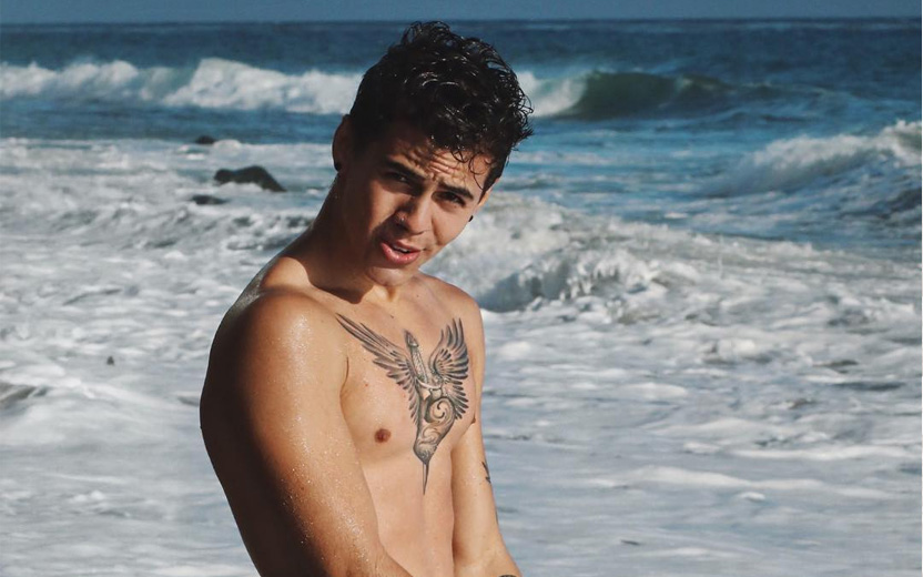 Biel sem camisa, com tatuagem de pássaro no peito em praia