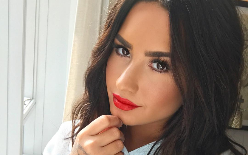 Demi Lovato com mão no queixo, usando batom vermelho e cabelos soltos e pretos