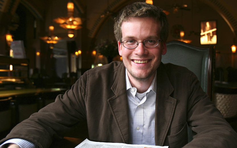 John Green sorri sentado em mesa de lugar que parece ser uma biblioteca. Ele usa camisa branca e casaco marrom