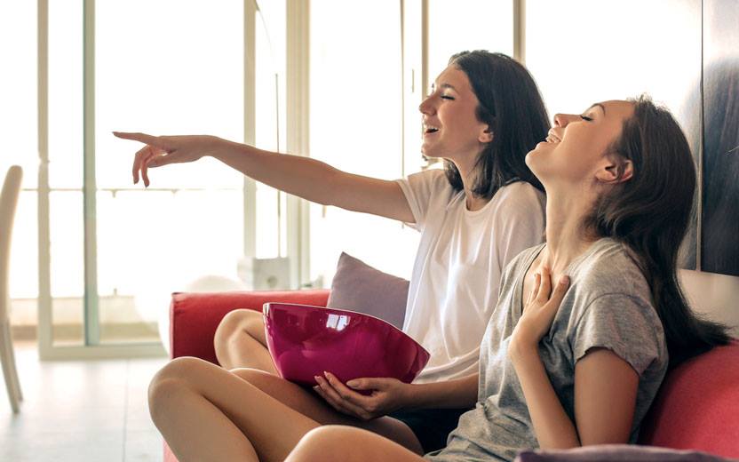 meninas rindo e apontando enquanto assistem tv sentadas em sofá e comendo pipoca