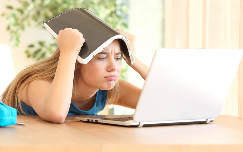 menina faz cara de cansada e coloca livro na cabeça enquanto olha tela de notebook