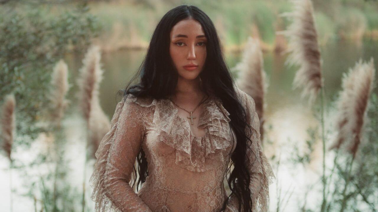 Noah Cyrus, filha de Billy Ray Cyrus, posa em frente a um lago usando um longo vestido de renda