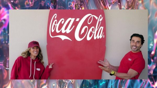 Karla e João Adibe Marques vestindo moletons vermelhos da Carmed posam sorridentes ao lado de uma grande placa com o logotipo da Coca-Cola.